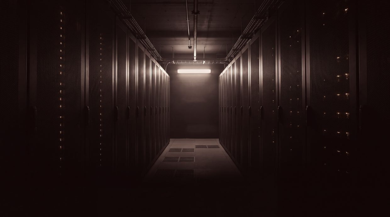 An aisle between server cabinets in an empty data hall at night, lit by a single overhead fixture. No person in the room.