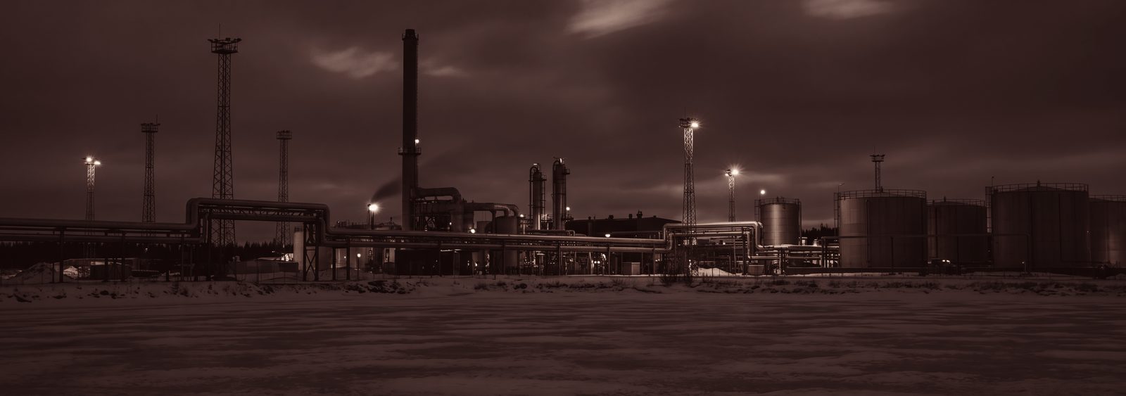 An industrial plant on a winter night: pipework, tanks and work lights above snow-covered ground. No people and no vehicles.